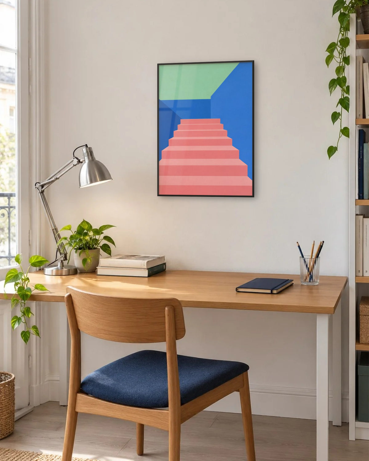 Affiche bureau escalier rose et bleu encadrée en noir au-dessus d'un bureau près d'une fenêtre