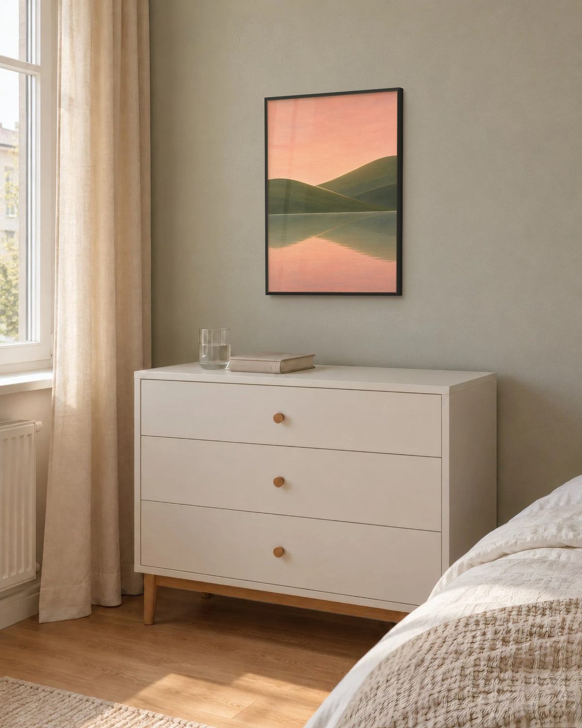 Affiche chambre lac rose et vert olive encadrée en noir sur un mur sauge, au-dessus d'une commode blanche