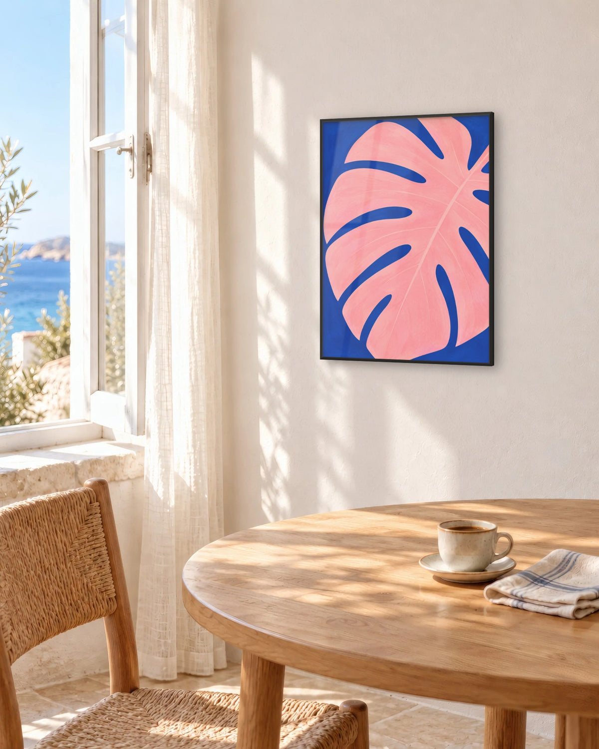 Affiche déco monstera rose et bleu cobalt encadrée en noir au-dessus d'une table face à la mer